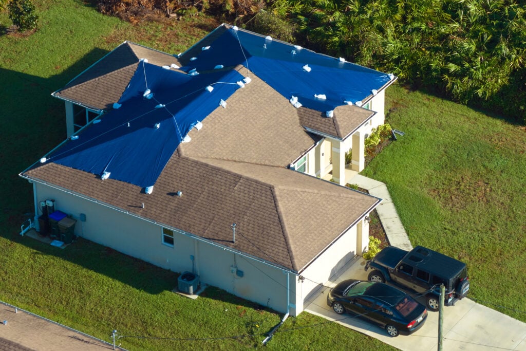 hail damage roof repair Aerial view of damaged in hurricane Ian house roof covered with blue protective tarp against rain water leaking until replacement of asphalt shingles