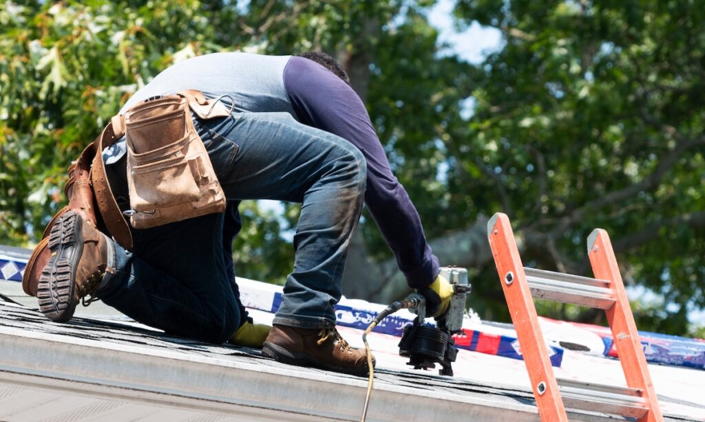 A contractor is kneeling on a residential roof using a nailgun to put new roof shingles on a house nest to a ladder.